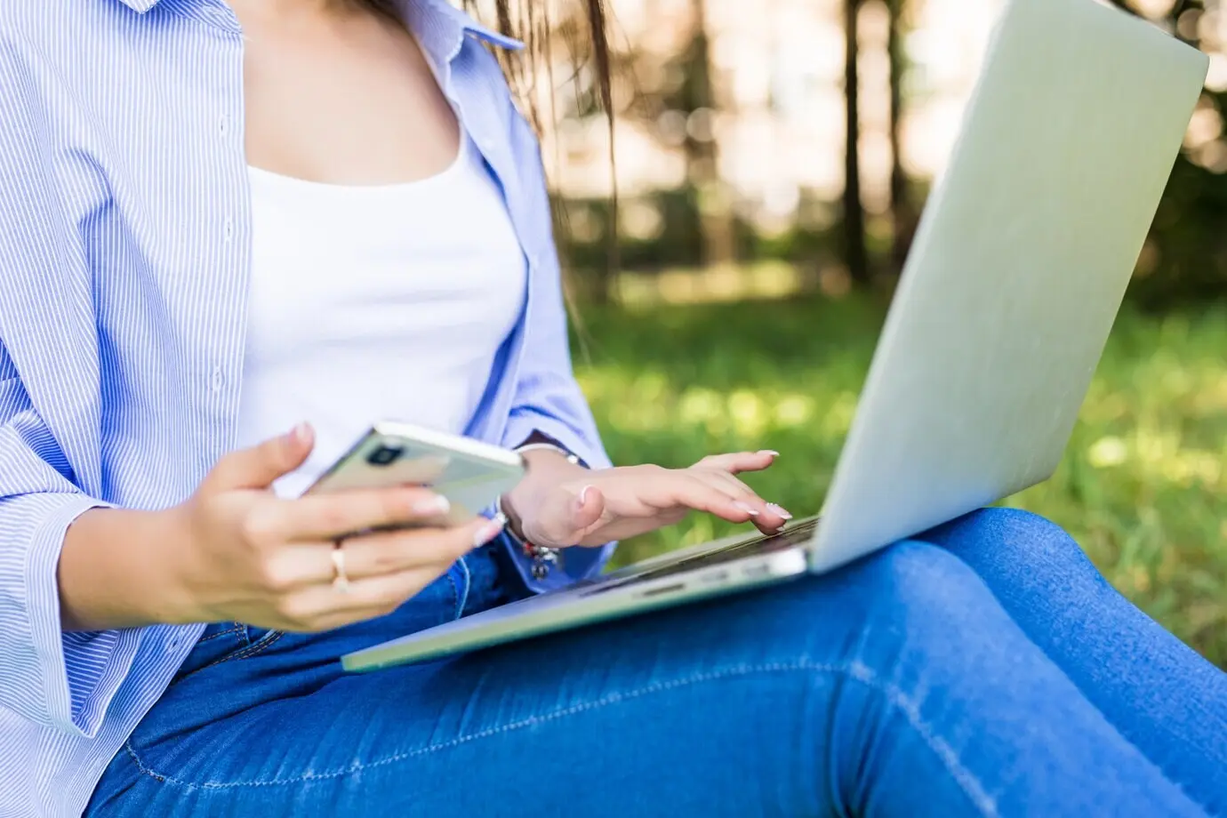 Красивая девушка в синих джинсах учится с ноутбуком и телефоном в городском парке.